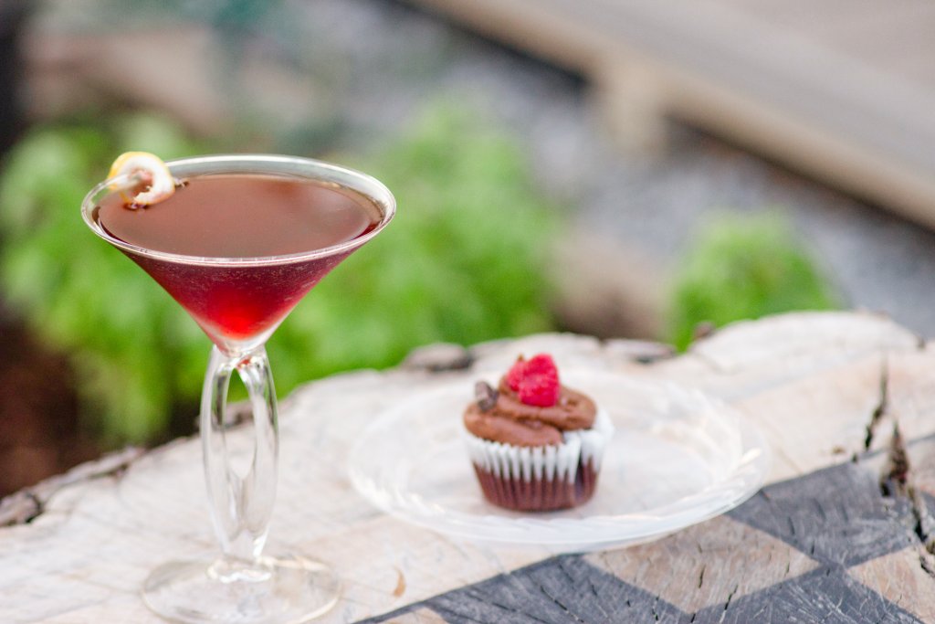 Events A vibrant red cocktail garnished with a lemon twist sits in a martini glass next to a chocolate cupcake topped with a single raspberry on a rustic wooden table in Williamson County, Tennessee.