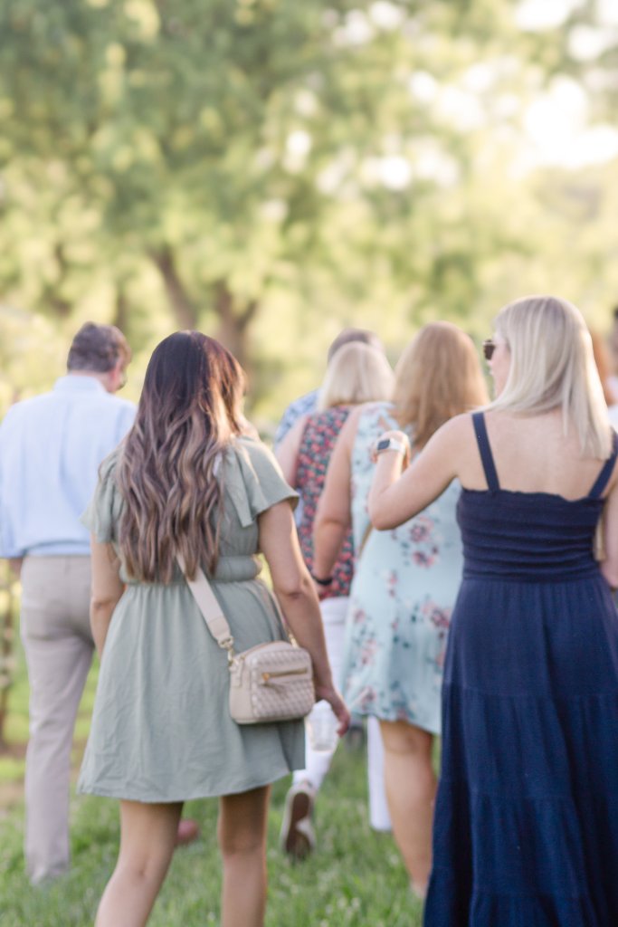 A group of women in summer dresses and men in casual attire stroll through a lush, green park on a sunny day in Williamson County, Tennessee.