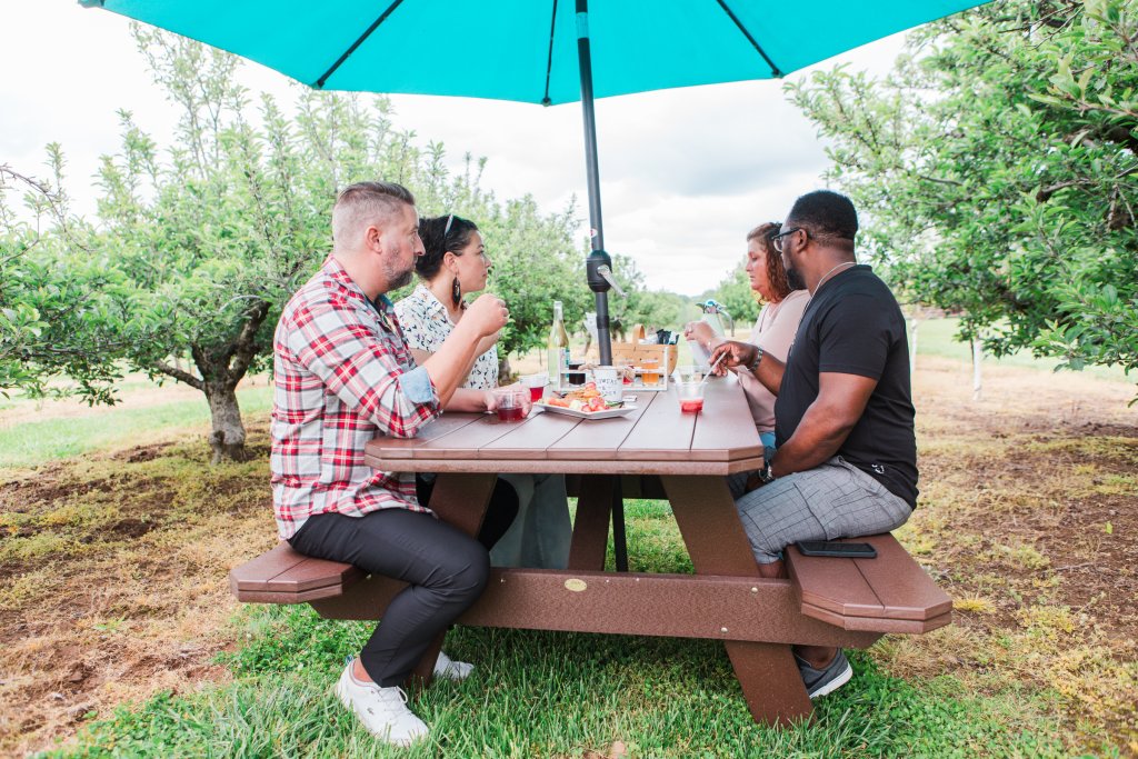 A group of four friends enjoys a picnic lunch under a blue umbrella at an apple orchard, surrounded by fruit trees in Williamson County, Tennessee.