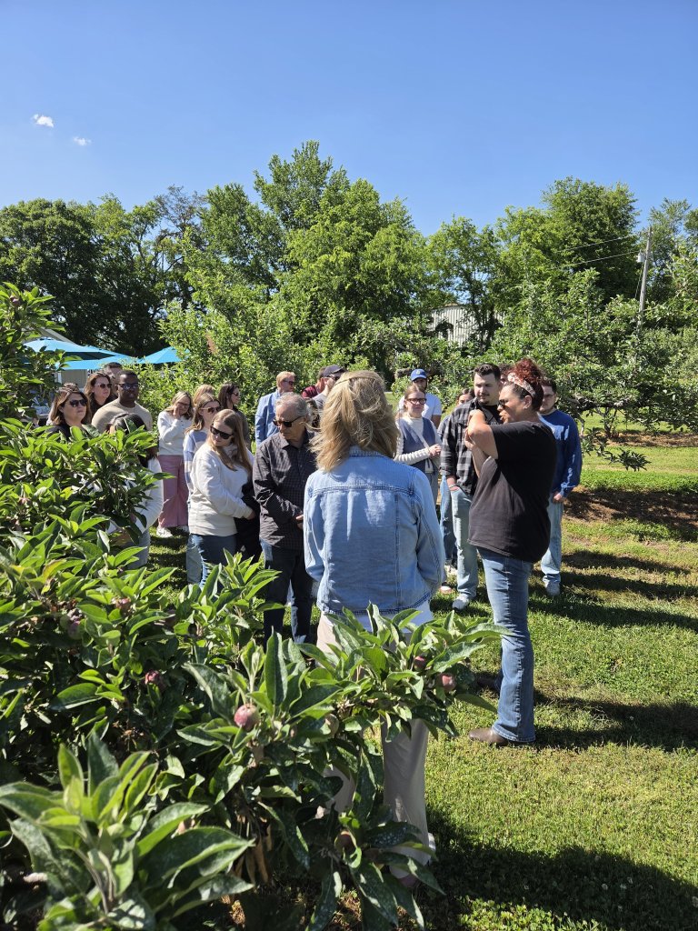 Events A group of people is gathered in an apple orchard, surrounded by lush green trees and ripe apples, under a clear blue sky in Williamson County, Tennessee.