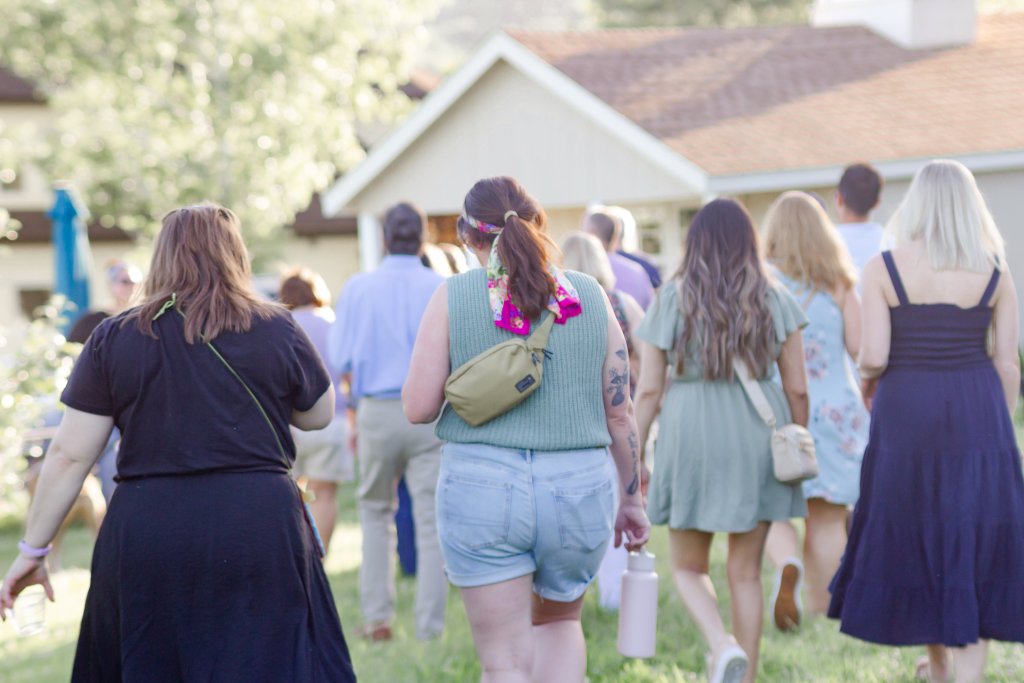 Taste & Tour A group of women in colorful summer dresses and men in casual attire walk together on a grassy lawn towards a white house with a brown roof in Williamson County, Tennessee.