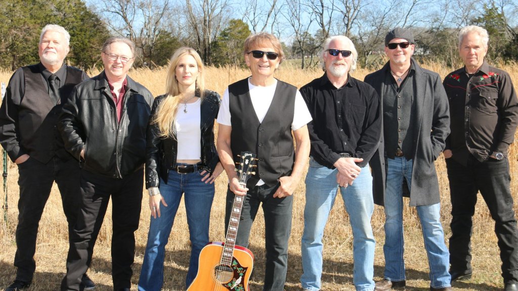 A group of seven musicians, dressed in black and blue, stand together outdoors with an acoustic guitar, surrounded by tall grass and trees in Williamson County, Tennessee.