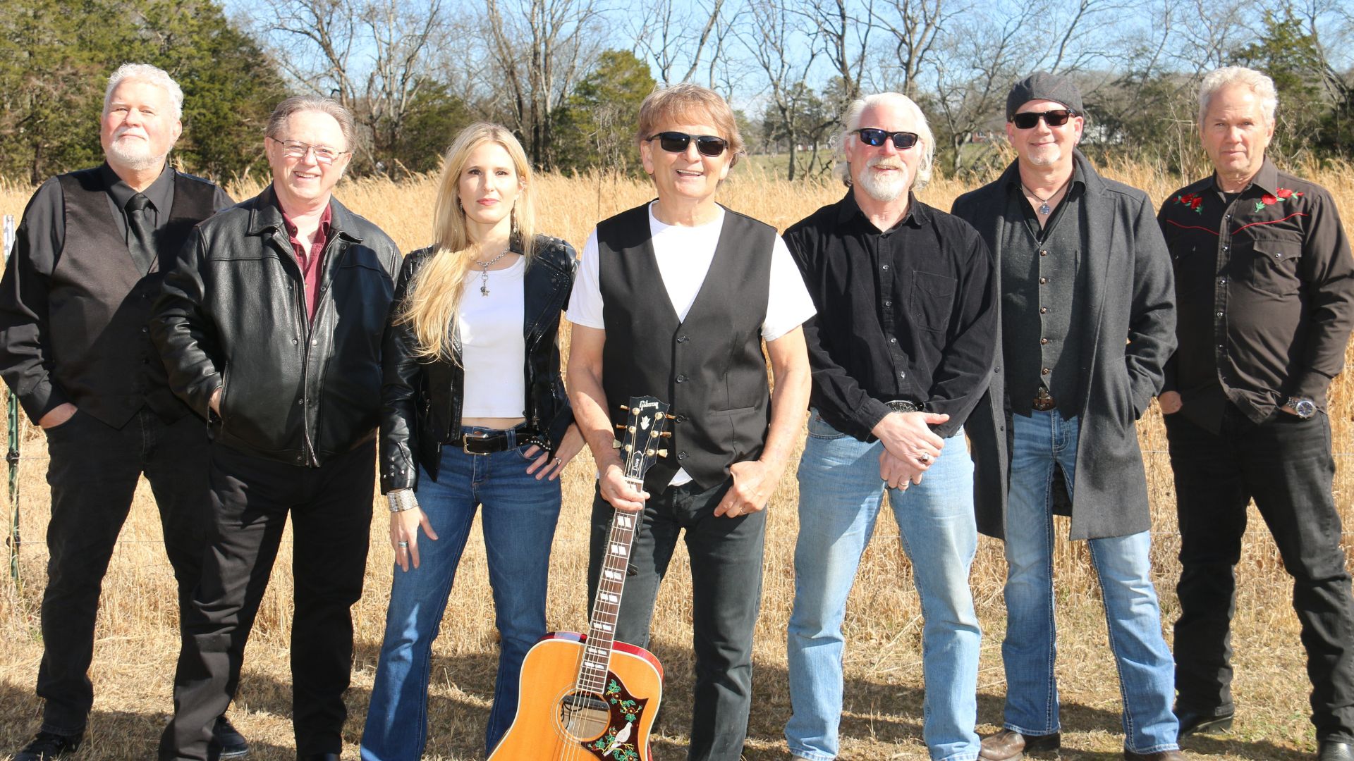 A group of seven musicians, dressed in black and blue, stand together outdoors with an acoustic guitar, surrounded by tall grass and trees in Williamson County, Tennessee.