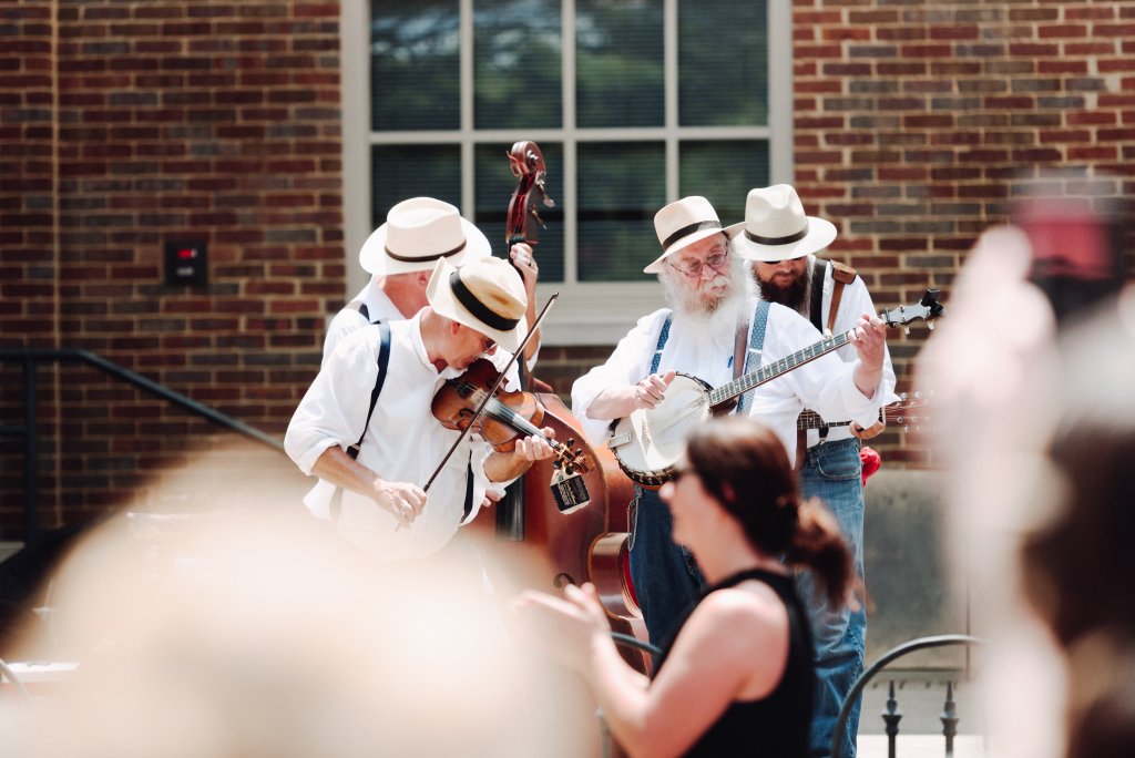 A lively string band performs outdoors, with five musicians dressed in white shirts, suspenders, and hats, playing violin, cello, banjo, and guitar in front of a brick building in Williamson County, Tennessee.