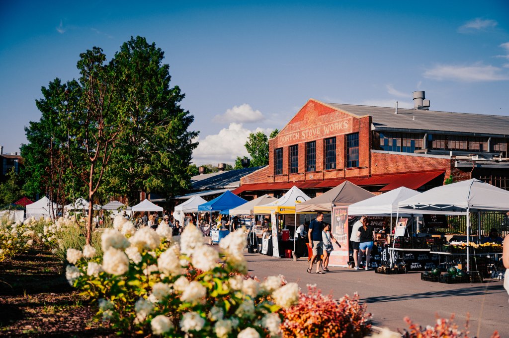 A bustling outdoor market with colorful tents and a historic red brick building in the background, surrounded by lush greenery and blooming white flowers in Williamson County, Tennessee.