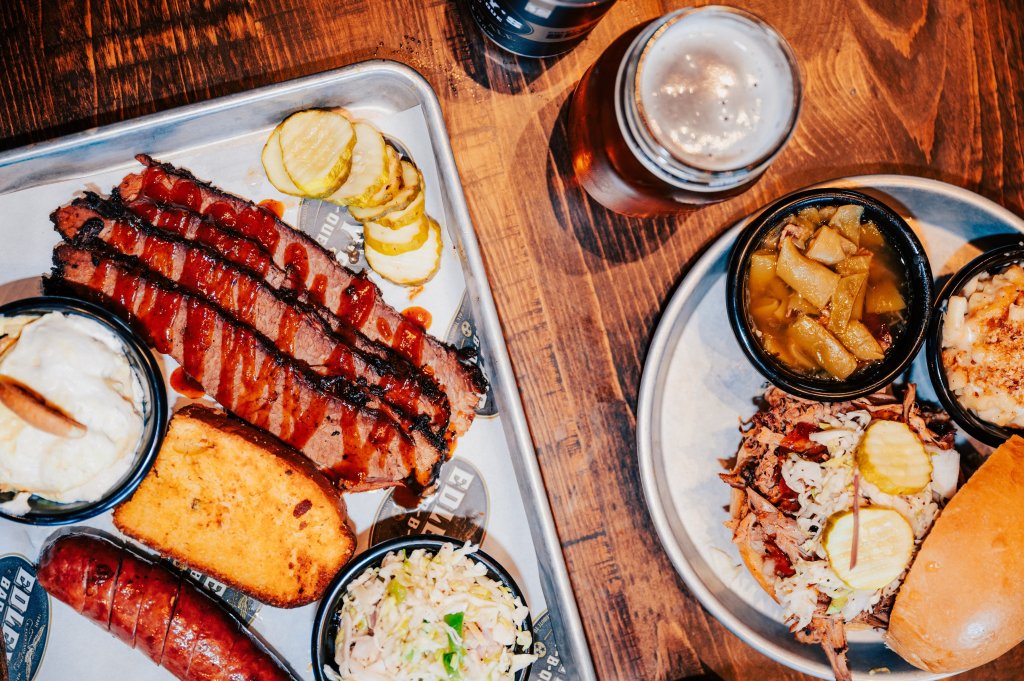 A hearty barbecue meal featuring smoked brisket, pulled pork sandwiches, and various sides including coleslaw, baked beans, and pickles, accompanied by a cold beer on a rustic wooden table in Williamson County, Tennessee.
