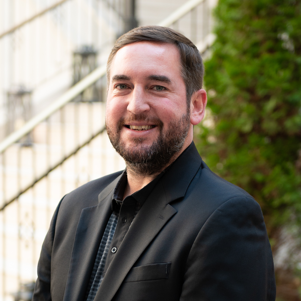 A man with a beard and mustache, wearing a black suit jacket over a black and white striped shirt, smiles warmly at the camera in Williamson County, Tennessee.