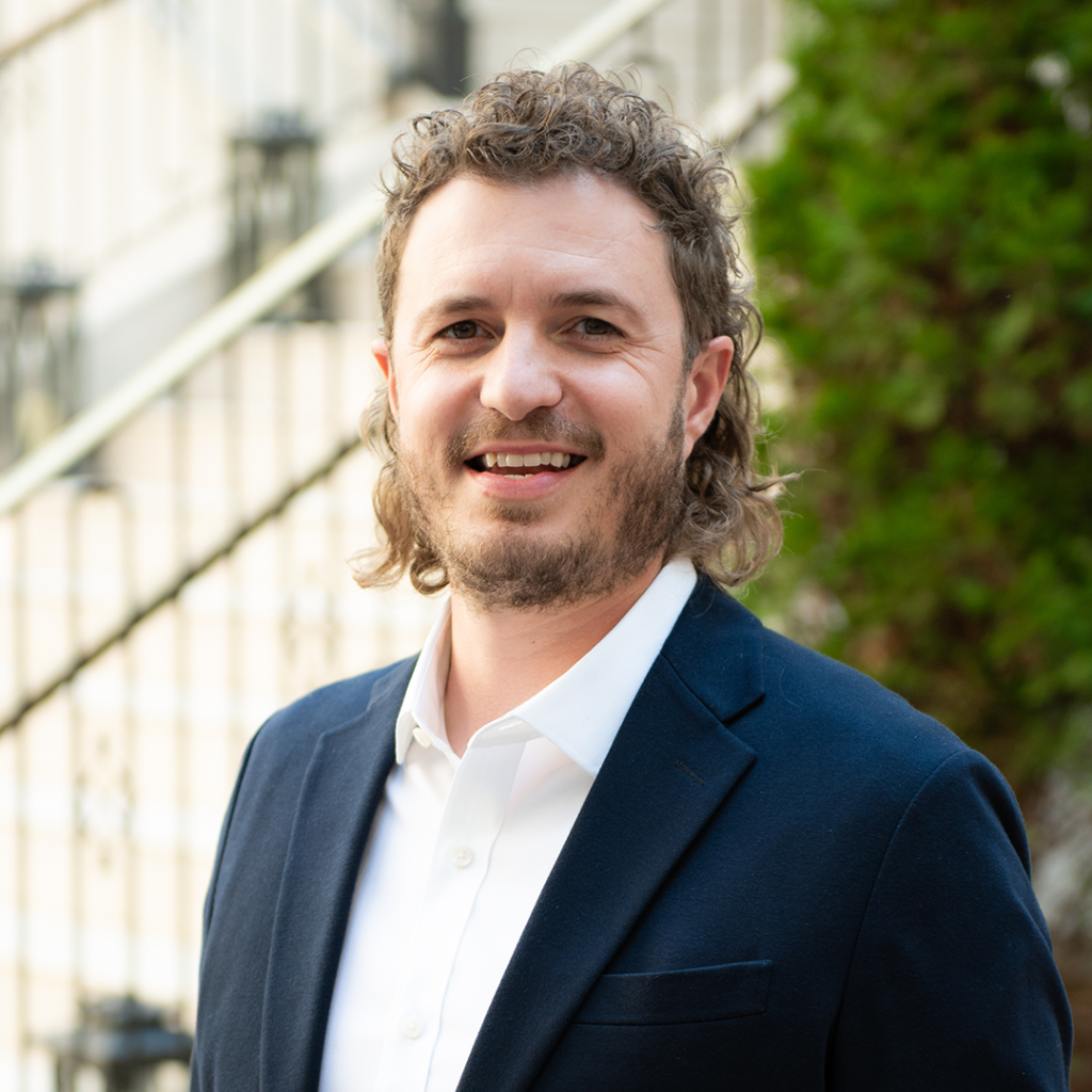 A man with curly hair and a beard, dressed in a dark blue suit jacket and white shirt, smiles warmly at the camera in Williamson County, Tennessee.