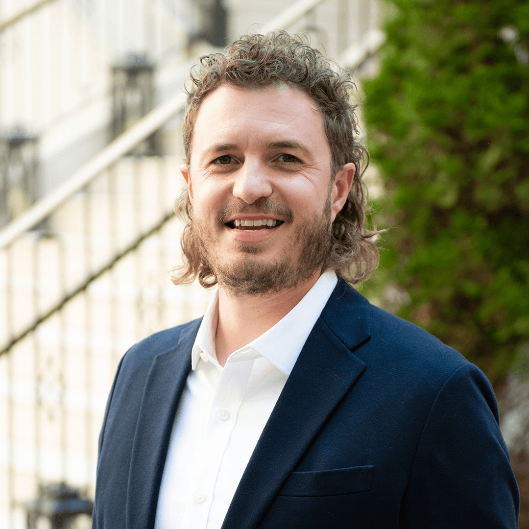 A man with curly hair and a beard, dressed in a dark blue suit jacket and white shirt, smiles warmly at the camera in Williamson County, Tennessee.