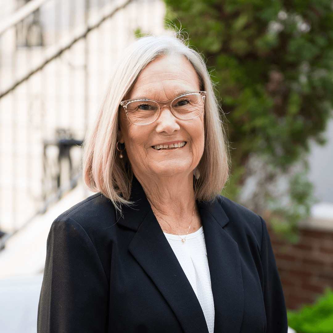 A smiling woman with short, blonde hair and glasses wearing a black blazer over a white blouse stands outdoors with greenery in the background in Williamson County, Tennessee.