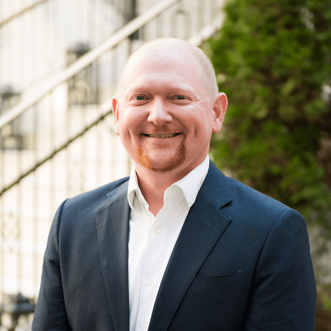 A bald man with a goatee smiles warmly, dressed in a dark blue suit and white shirt, standing in front of a white railing and green shrubbery in Williamson County, Tennessee.