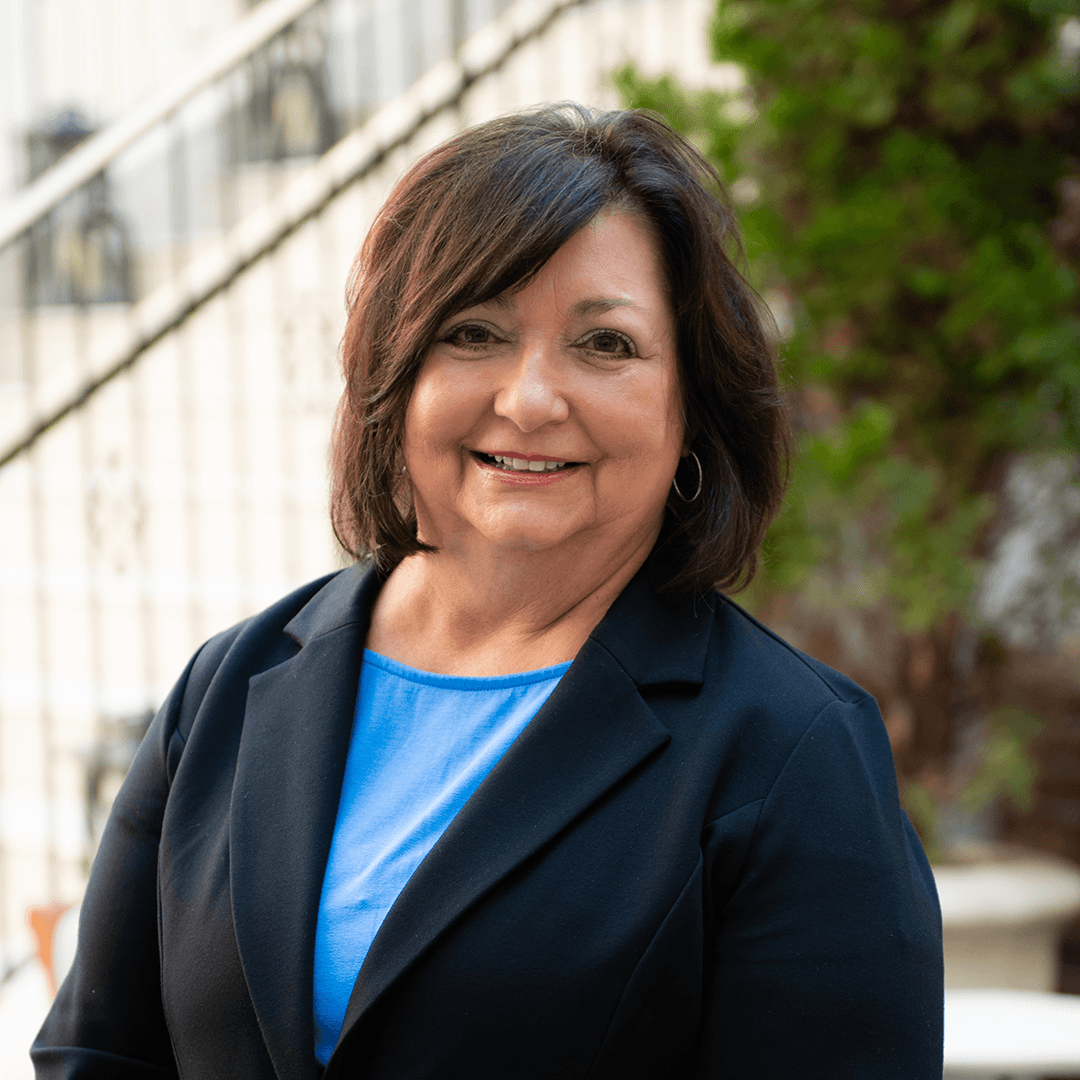 A woman with shoulder-length dark hair smiles warmly, wearing a black blazer over a blue top, standing in front of a staircase with greenery in the background in Williamson County, Tennessee.