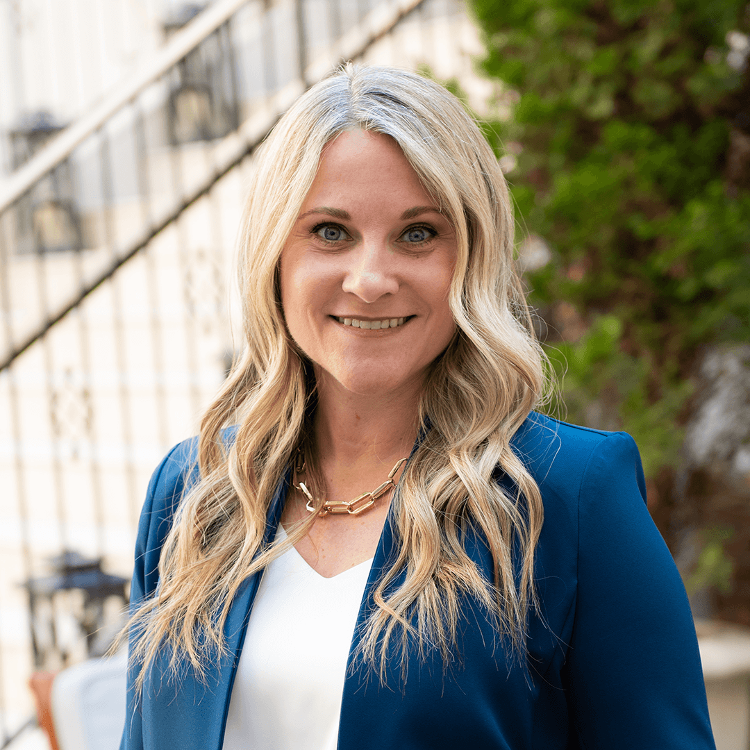 A woman with long, wavy blonde hair is smiling warmly at the camera, wearing a stylish blue blazer over a white top, accessorized with a gold chain necklace in Williamson County, Tennessee.