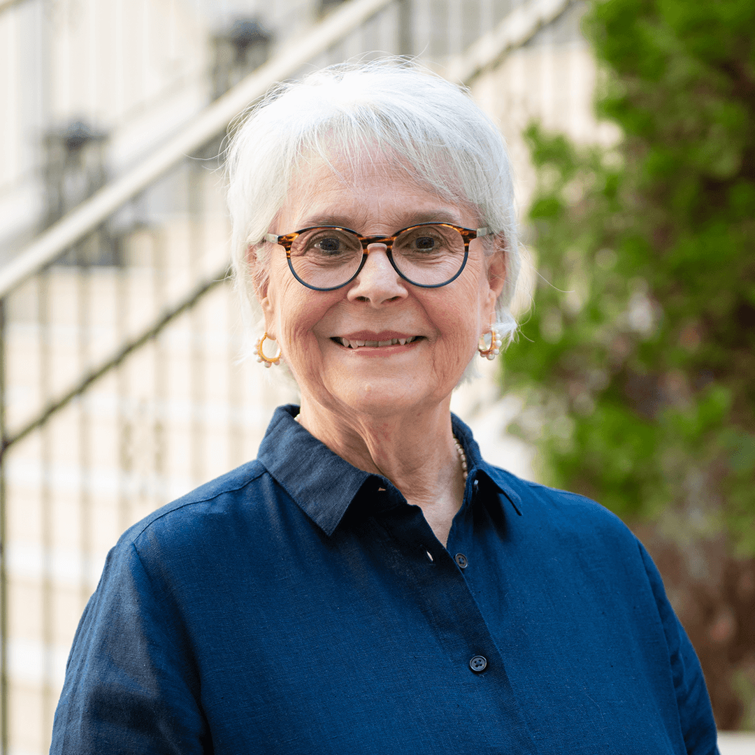 An elderly woman with short white hair, wearing glasses, a dark blue button-up shirt, and pearl earrings, smiles warmly at the camera in Williamson County, Tennessee.