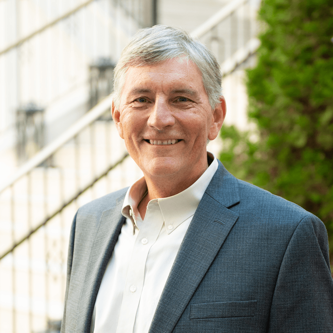 A man with gray hair smiles warmly while wearing a gray suit jacket over a white shirt, standing outdoors with a staircase and greenery in the background in Williamson County, Tennessee.