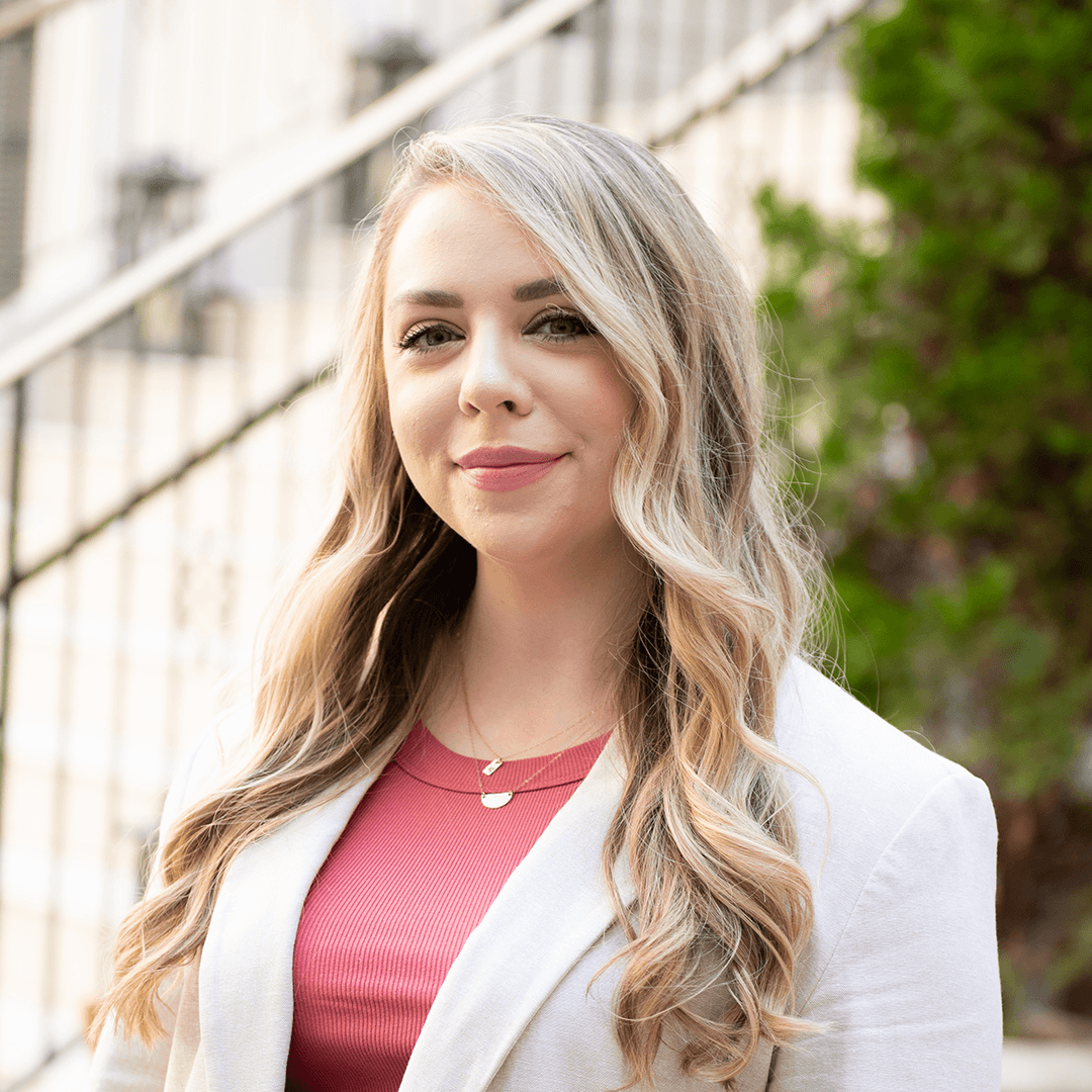 A young woman with long, wavy blonde hair smiles warmly at the camera, wearing a stylish white blazer over a pink top in Williamson County, Tennessee.