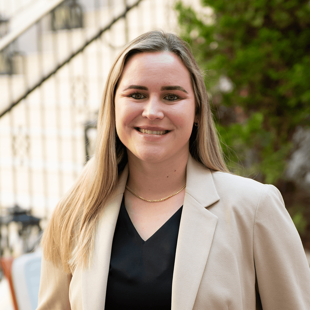 A young woman with long blonde hair smiles confidently, wearing a beige blazer over a black top, standing in front of a white building with a staircase and greenery in the background in Williamson County, Tennessee.