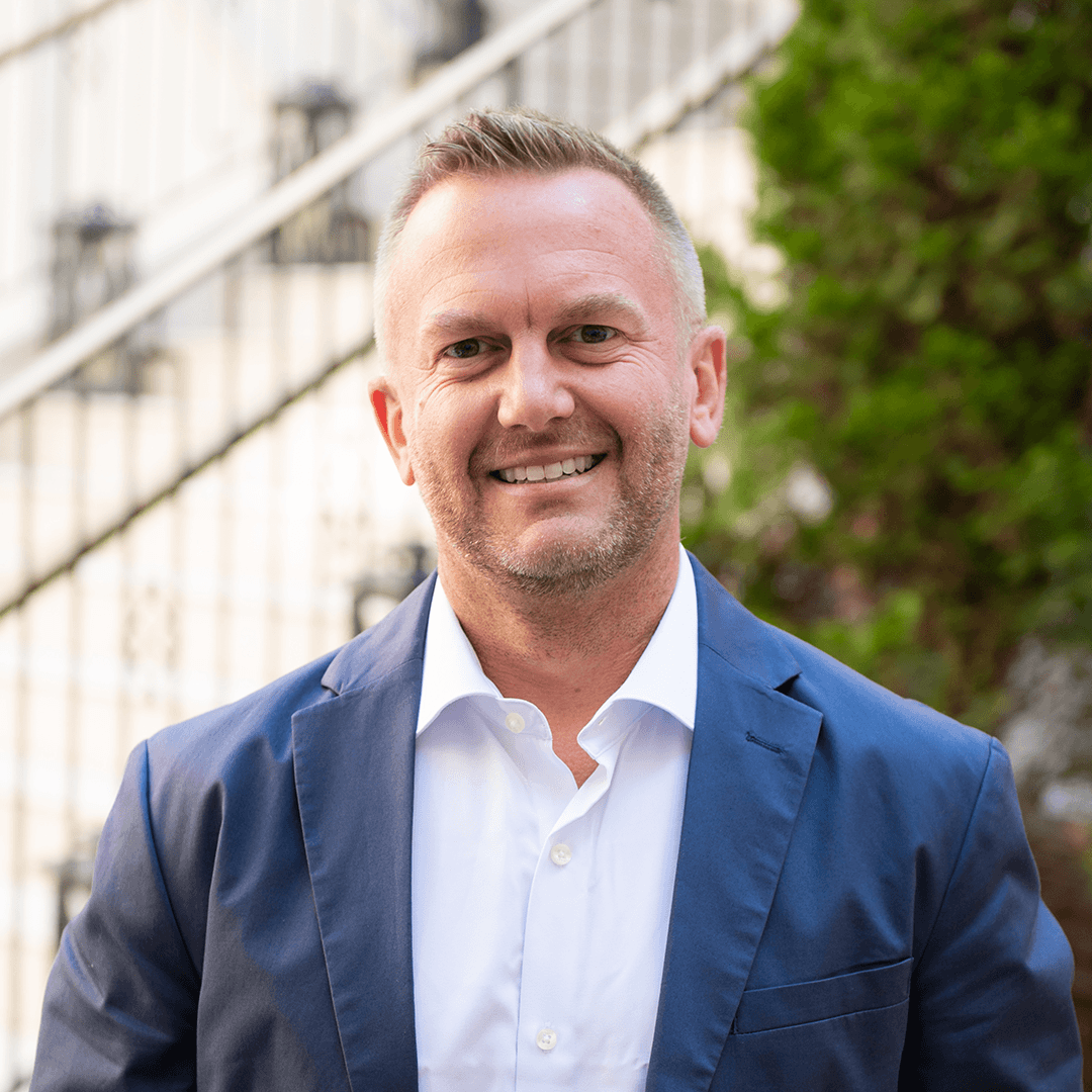 A man in a navy blue suit jacket and white shirt stands outdoors with a smile, with a blurred background featuring greenery and a staircase in Williamson County, Tennessee.