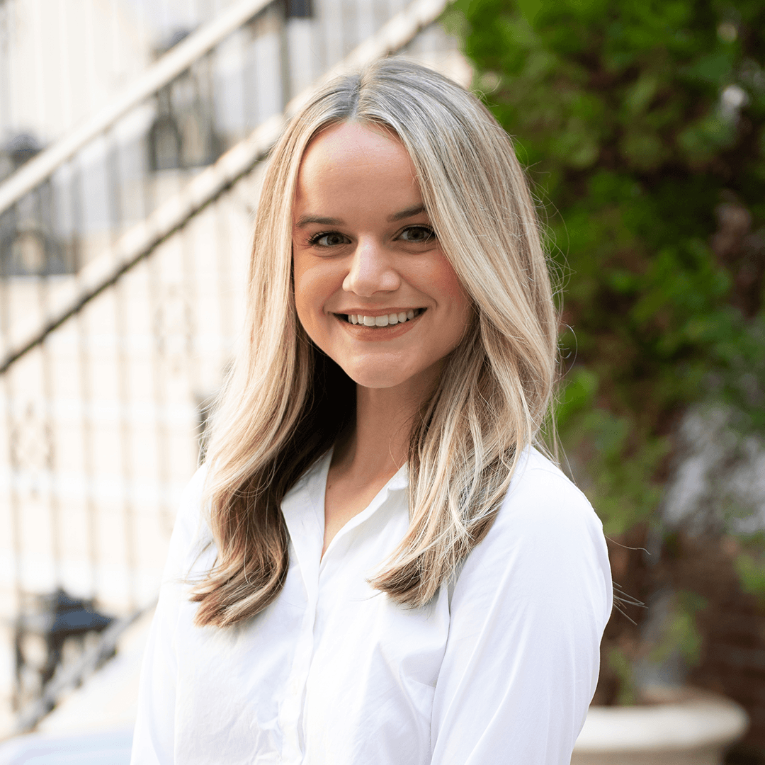 A young woman with long blonde hair smiles brightly, standing in front of a white building with a staircase in Williamson County, Tennessee.