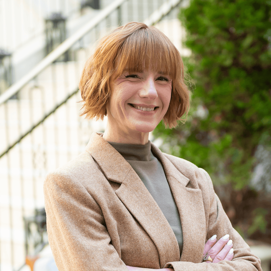 A woman with short, wavy red hair smiles warmly, wearing a beige blazer over a gray turtleneck, standing in front of a white railing and green foliage in Williamson County, Tennessee.