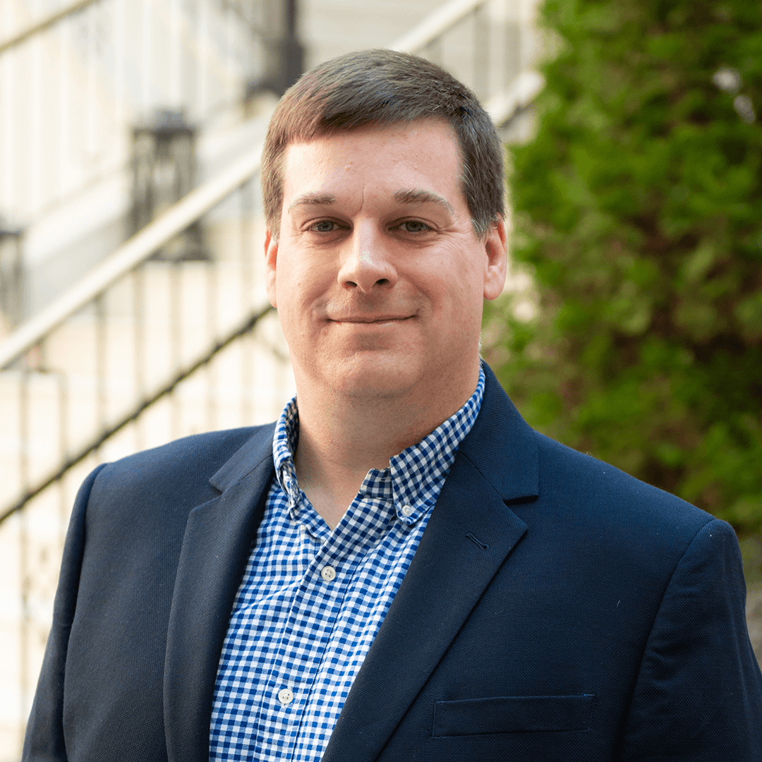 A man wearing a navy blue blazer over a blue and white checkered shirt, standing outdoors with a staircase and greenery in the background in Williamson County, Tennessee.