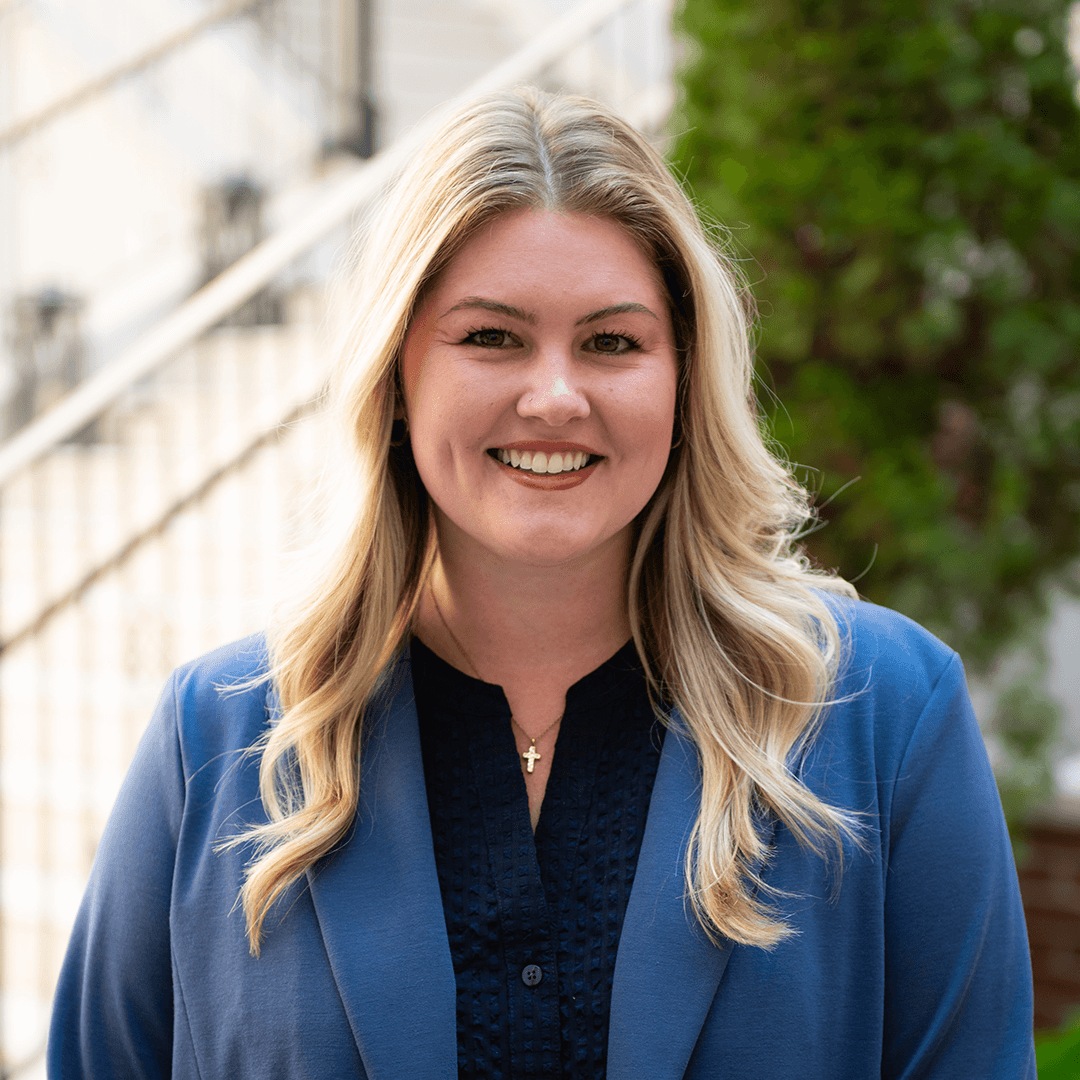 A smiling blonde woman wearing a blue blazer stands in front of a white railing and green foliage in Williamson County, Tennessee.