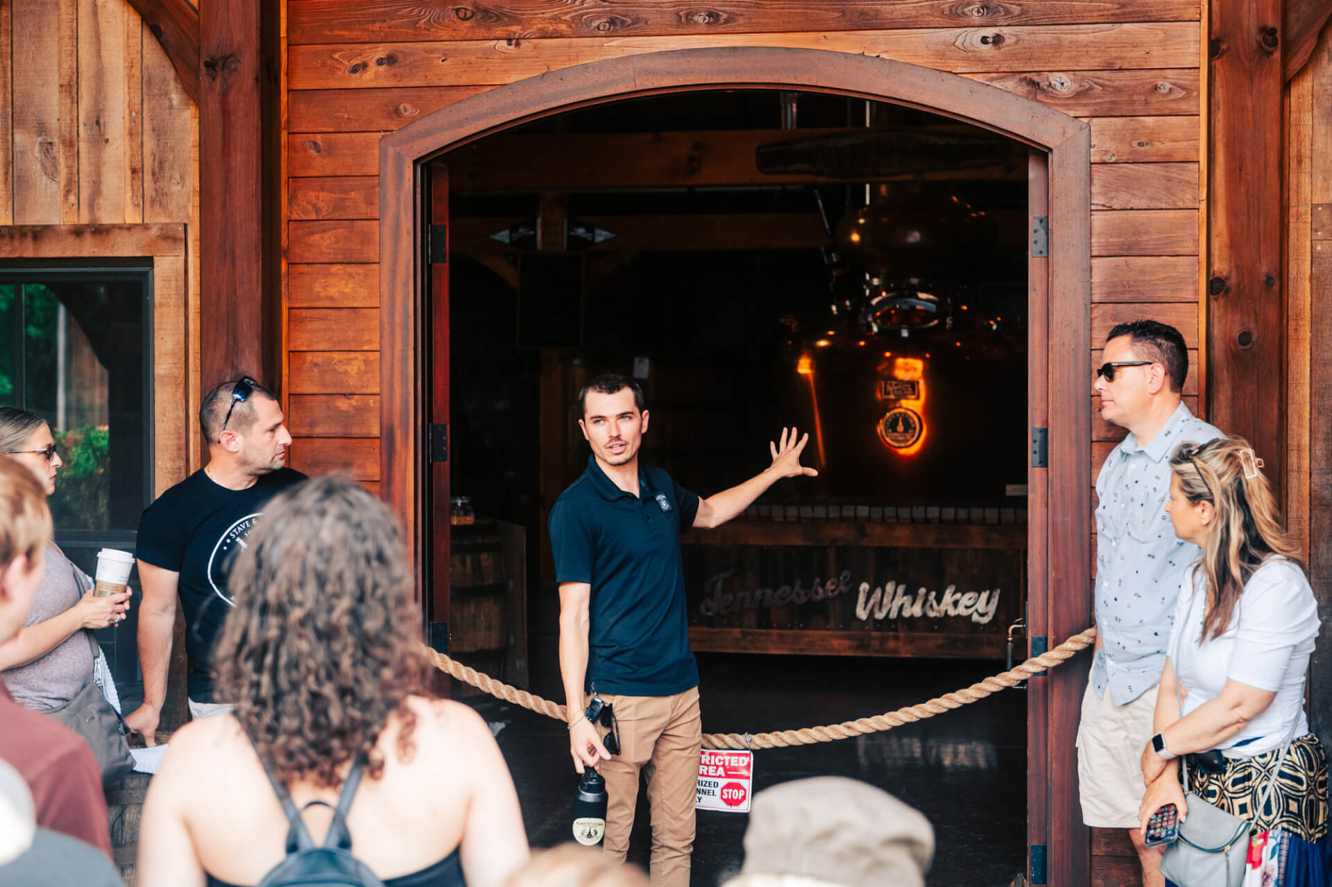 A group of people gather outside a wooden building with a large archway, where a man in a dark polo shirt gestures towards the interior, which is dimly lit and features a large copper still in Williamson County, Tennessee.