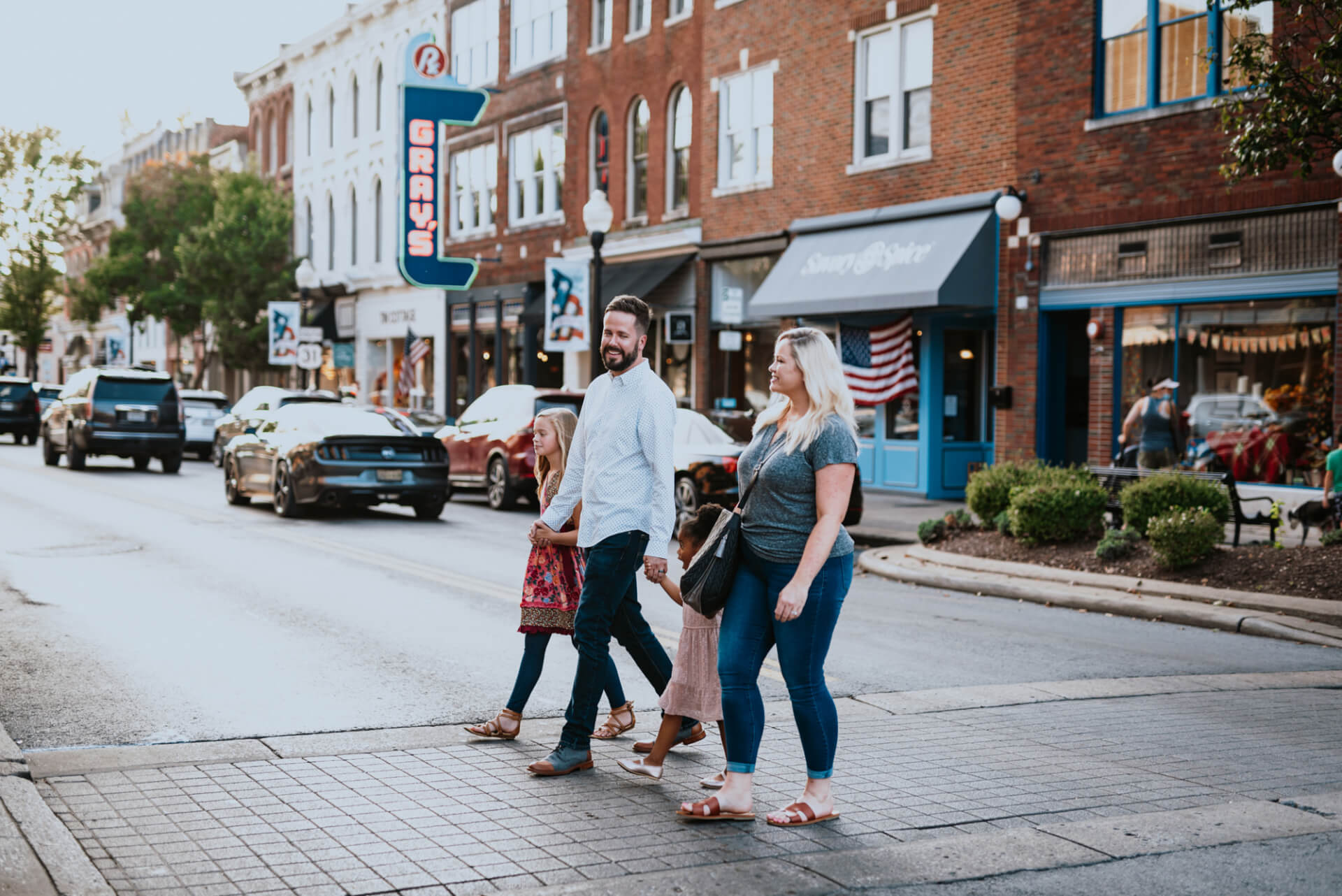 A family of four walks hand-in-hand across a quaint town square, surrounded by historic brick buildings and American flags in Williamson County, Tennessee.