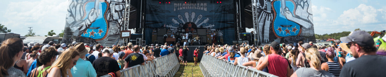 A large crowd of people gathered in front of a stage with colorful murals, enjoying an outdoor music festival on a sunny day in Williamson County, Tennessee.