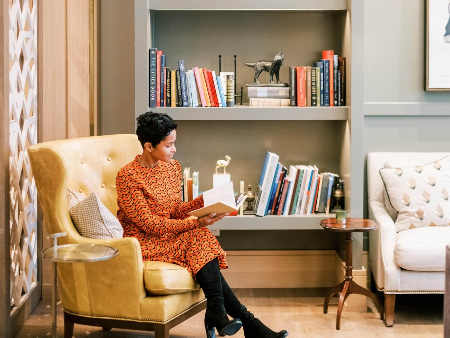 A woman in an orange patterned dress sits in a yellow armchair, engrossed in a book, with a well-stocked bookshelf and a cozy living room setting behind her in Williamson County, Tennessee.