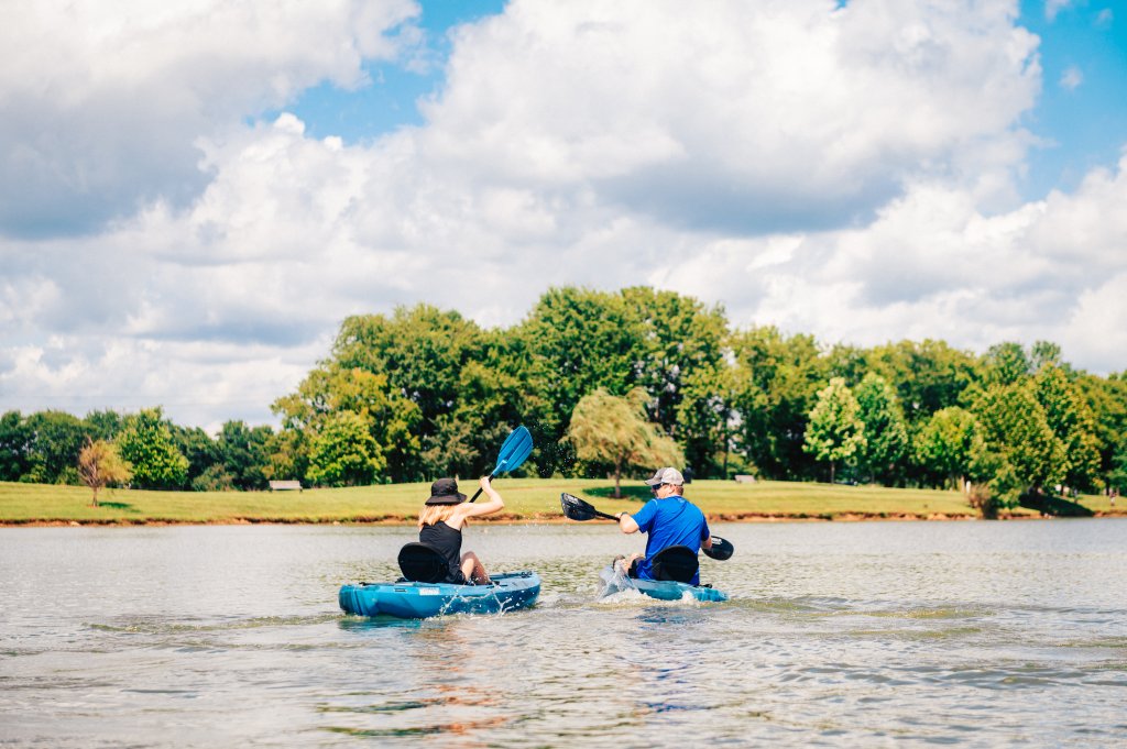 Two kayakers paddle across a serene lake surrounded by lush green trees and a grassy shore under a partly cloudy sky in Williamson County, Tennessee.