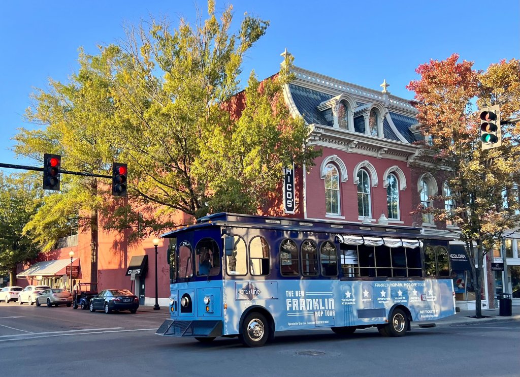 End of Summer Escape A blue trolley bus with 'The New Franklin Hop-On Hop-Off Tour' painted on its side travels down a city street lined with colorful autumn trees and historic buildings in Williamson County, Tennessee.