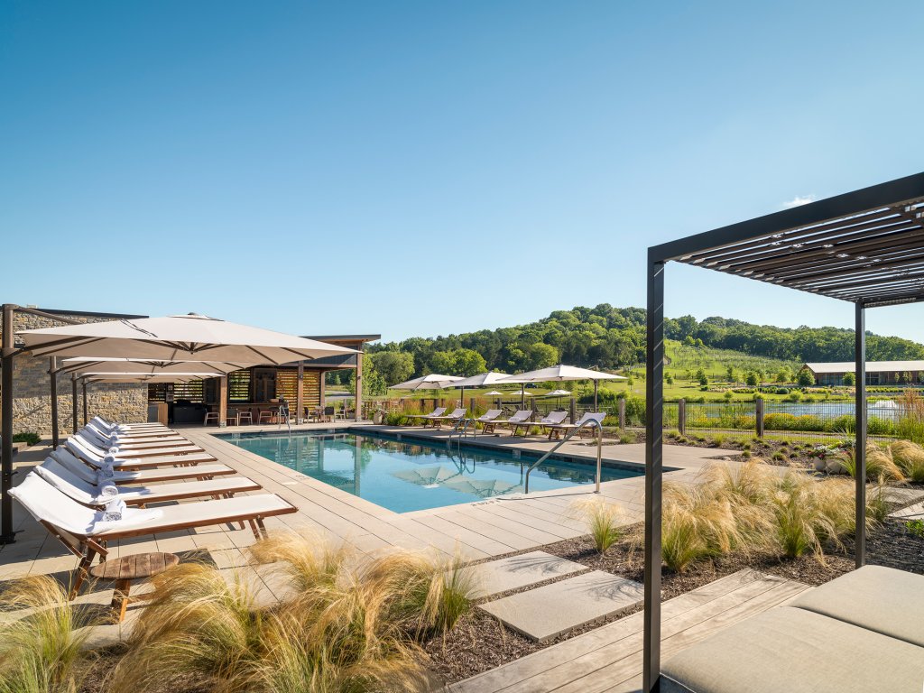 End of Summer Escape A serene outdoor swimming pool area with multiple lounge chairs, umbrellas, and a modern pergola structure, set against a backdrop of lush green hills and a clear blue sky in Williamson County, Tennessee.