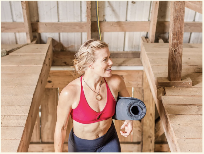 Yoga in the Barn A woman in a red sports bra and black leggings smiles while holding a yoga mat in a rustic wooden barn in Williamson County, Tennessee.