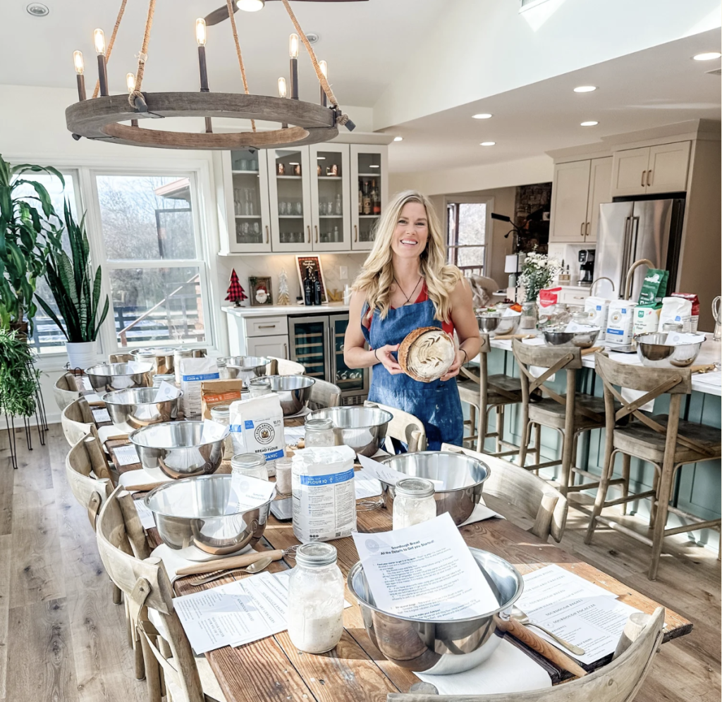 Events A woman in an apron stands in a bright kitchen filled with tables set for a bread-making class, holding a freshly baked loaf in Williamson County, Tennessee.