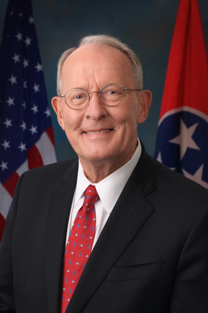 A smiling man in a black suit with a red tie stands between the American and Tennessee state flags in Williamson County, Tennessee.