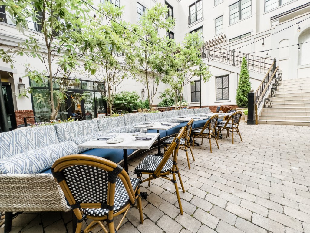 An outdoor dining area with a long blue and white striped sofa and matching chairs, surrounded by greenery and a white building with black shutters in Williamson County, Tennessee.