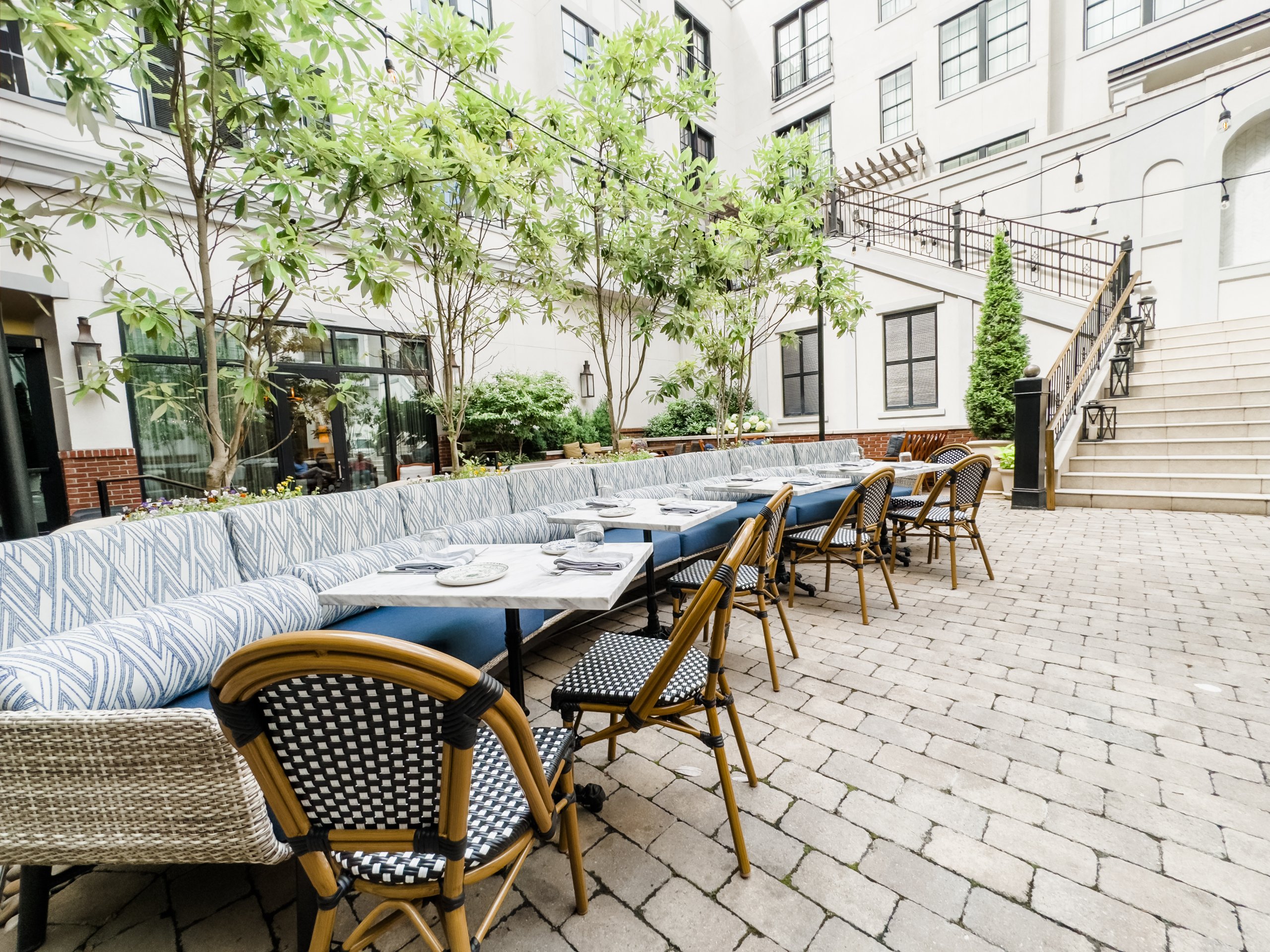 The Harpeth Hotel Sound Bath An outdoor dining area with a long blue and white striped sofa and matching chairs, surrounded by greenery and a white building with black shutters in Williamson County, Tennessee.