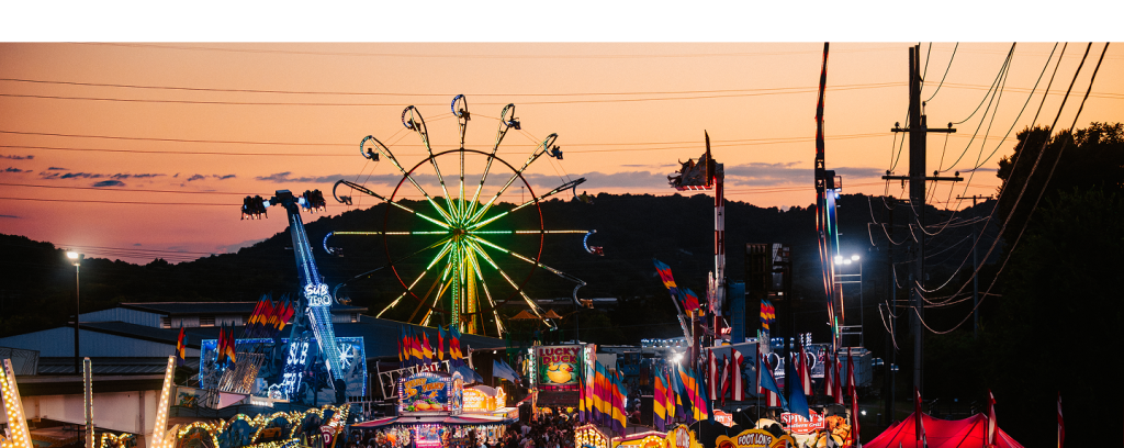 A vibrant carnival at sunset with a towering Ferris wheel, colorful rides, and festive flags against a backdrop of rolling hills in Williamson County, Tennessee.