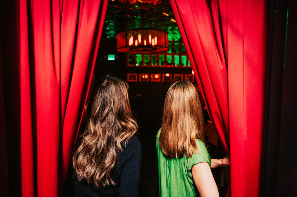 The 8 Best Late-Night Spots for Live Music, Cocktails & More in Franklin Two women with long hair stand behind red curtains, looking into a dimly lit room with a chandelier and green lighting in Williamson County, Tennessee.