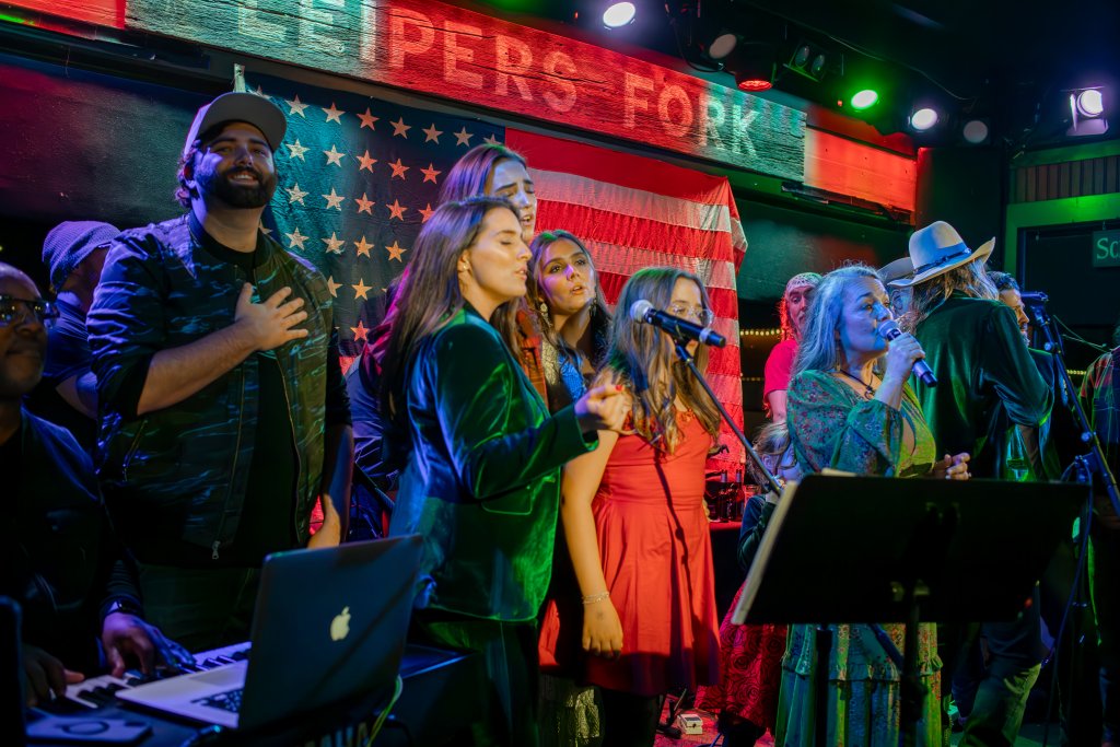 The 8 Best Late-Night Spots for Live Music, Cocktails & More in Franklin A lively band performs on stage, with a diverse group of musicians singing and playing instruments under a backdrop of an American flag and neon signs in Williamson County, Tennessee.