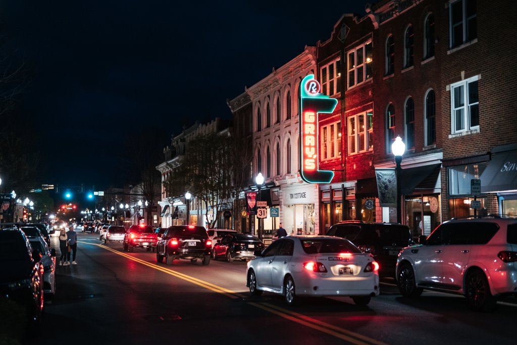 The 8 Best Late-Night Spots for Live Music, Cocktails & More in Franklin A bustling downtown street at night, illuminated by neon signs and streetlights, with cars driving and pedestrians walking in Williamson County, Tennessee.