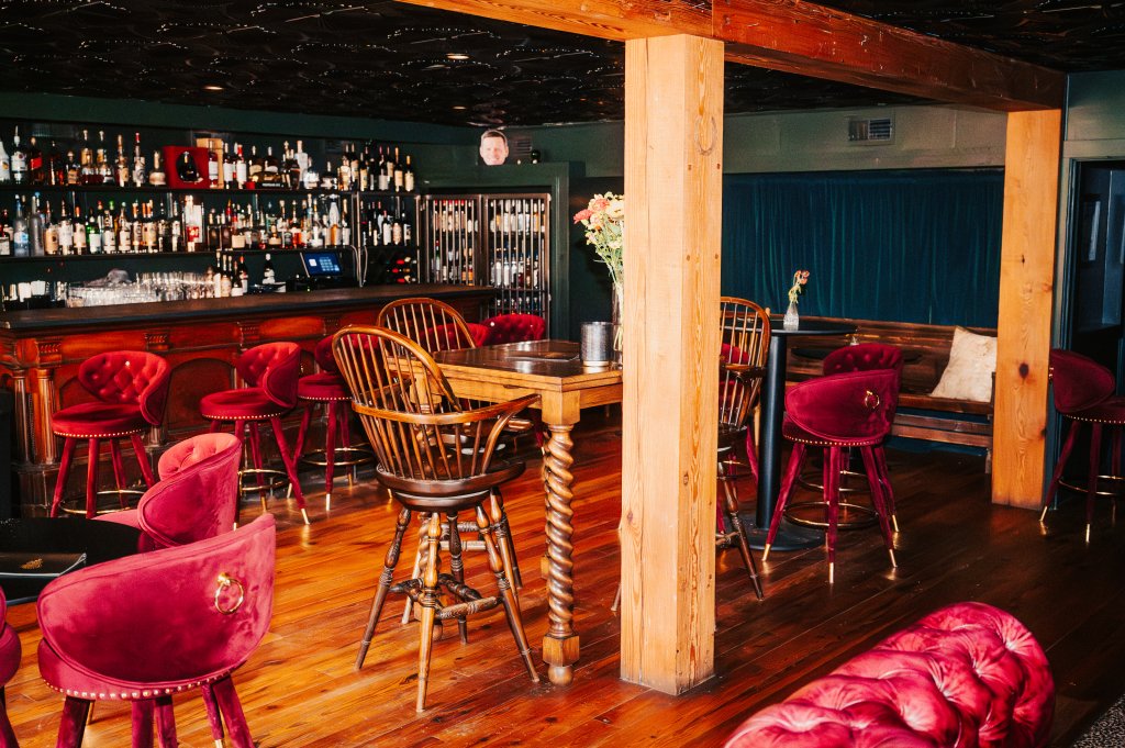 The 8 Best Late-Night Spots for Live Music, Cocktails & More in Franklin A dimly lit, cozy bar with a rustic wooden interior, featuring a variety of plush red velvet chairs and stools, a central wooden table, and a well-stocked bar area with shelves of liquor bottles in Williamson County, Tennessee.