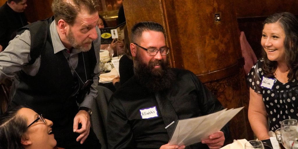 Events A lively group of people, including a bearded man in a black suit, a woman in a polka-dotted dress, and a man in a vest, are engaged in conversation and laughter at a dinner table in Williamson County, Tennessee.
