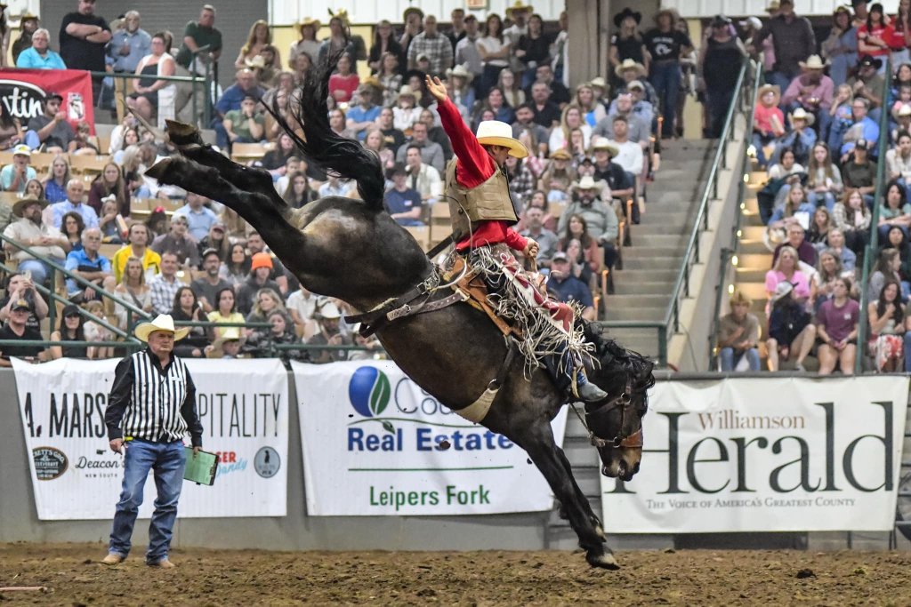 Events A cowboy in a red shirt and white cowboy hat is riding a bucking bronco in a rodeo arena, with a crowd of spectators watching from the stands in Williamson County, Tennessee.