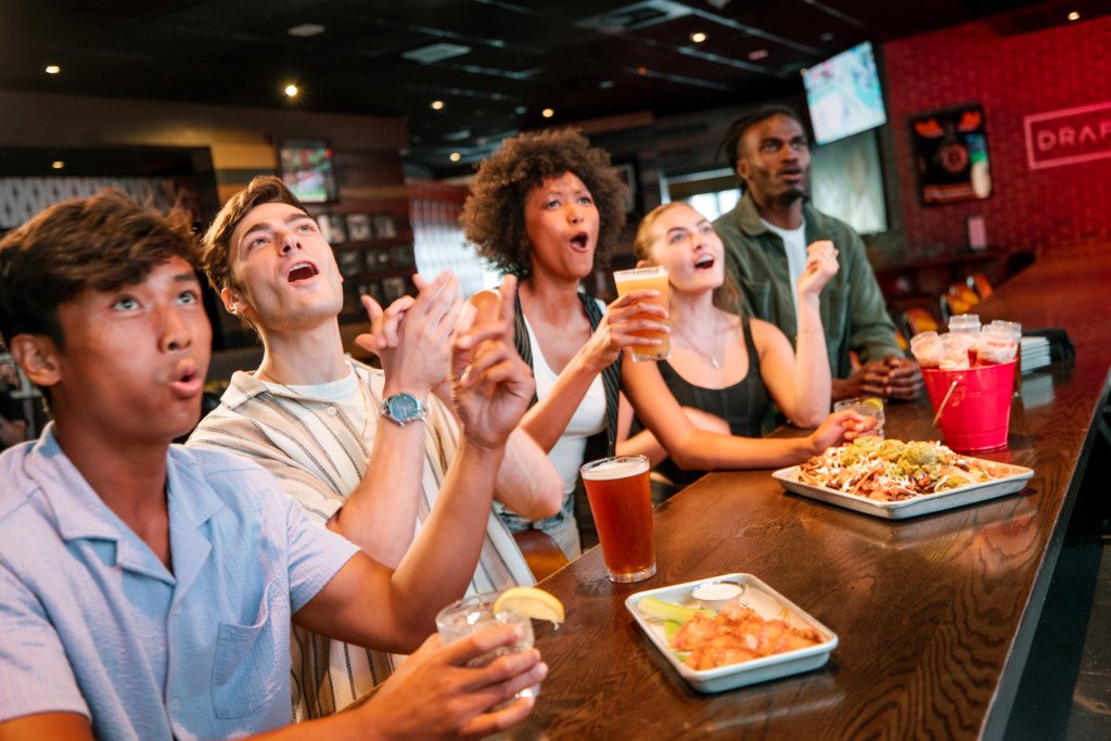 World Tournament Watch Parties at Kings A group of five friends, three men and two women, are gathered around a bar table, cheering and clapping while watching a sports game on a TV screen above them. They are enjoying various drinks and snacks, including beer, margaritas, and plates of nachos and chicken wings in Williamson County, Tennessee.