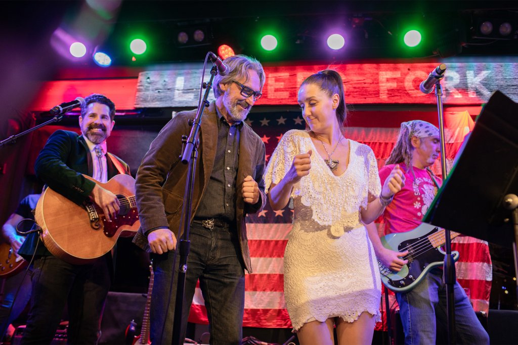 Things To Do A lively band performs on stage, featuring a man in a brown jacket playing an acoustic guitar, a woman in a white dress singing into a microphone, and two other musicians with electric guitars, all set against a backdrop of American flags and colorful stage lights in Williamson County, Tennessee.