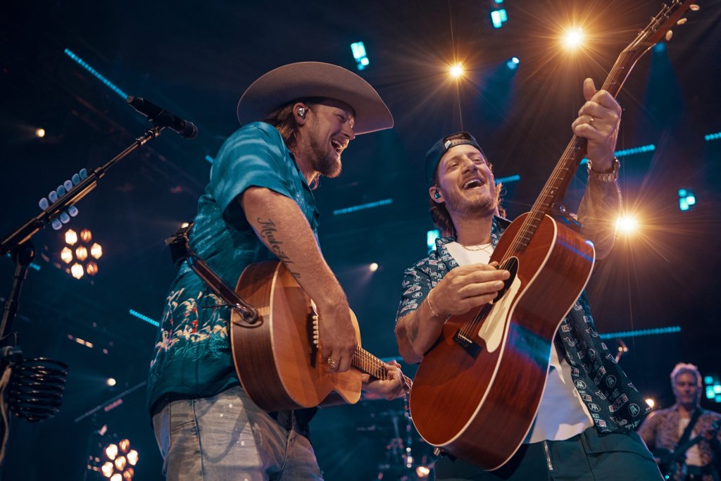 Two musicians, one wearing a cowboy hat and the other a baseball cap, play acoustic guitars on stage with joyful expressions in Williamson County, Tennessee.