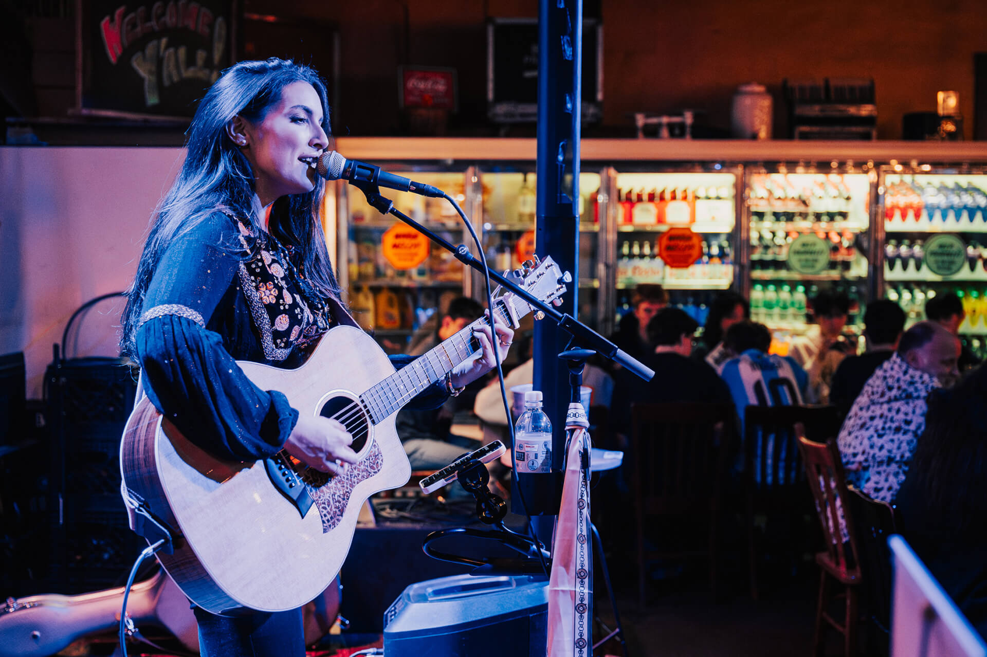 Where to Stay A woman with long dark hair performs an acoustic set in a dimly lit bar, singing into a microphone while playing an acoustic guitar in Williamson County, Tennessee.