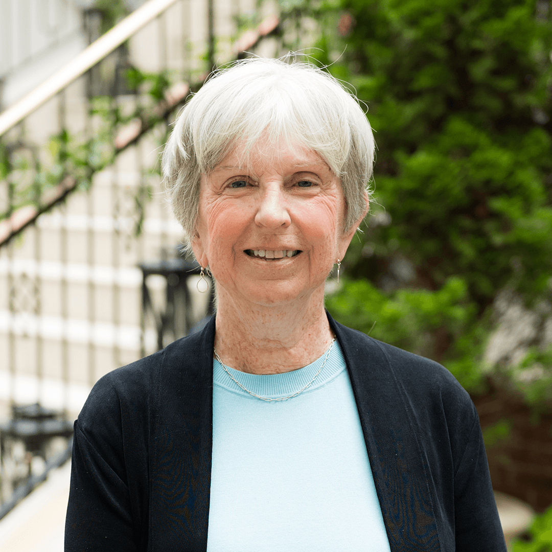 Meet The Team A smiling woman with short white hair wearing a black cardigan over a light blue shirt stands in front of a staircase with greenery in the background in Williamson County, Tennessee.